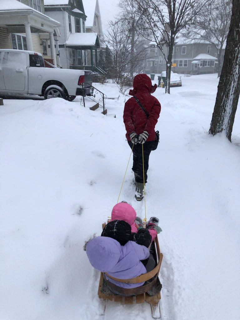 Car lite: an adult pulls three kids on a sled down a snowy sidewalk