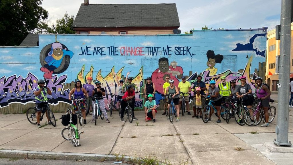 Group bike ride photo; "we are the change that we seek" mural.