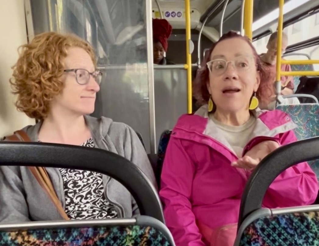 Two women sitting next to each other on a bus. One is talking while the other listens