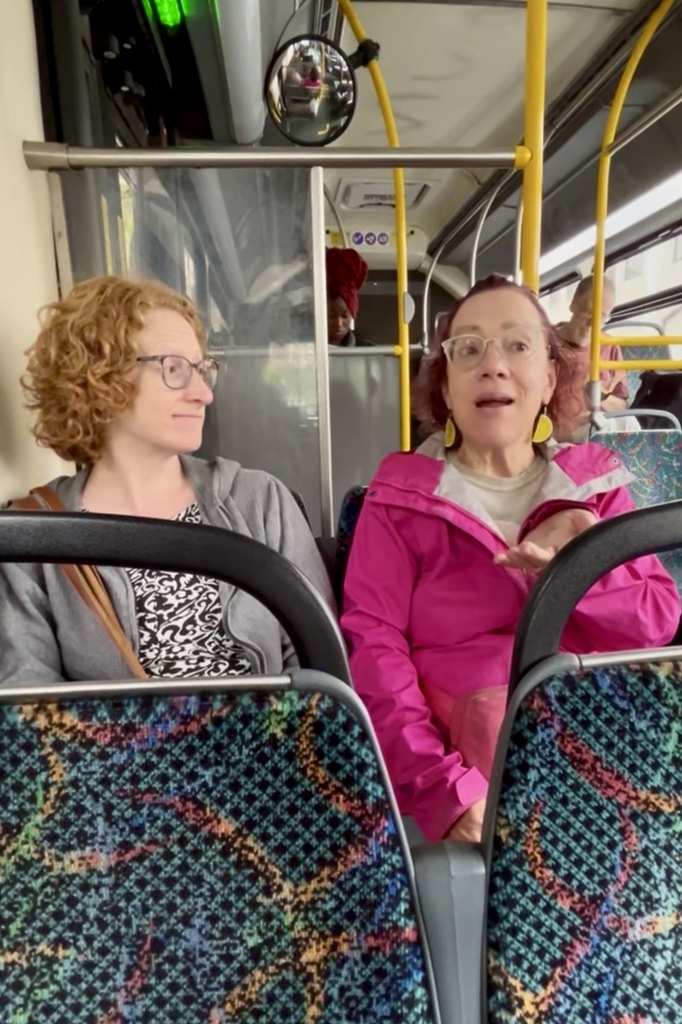 Two women sitting next to each other on a bus. One is talking while the other listens